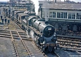 A last moment of glory for Standard Class 4 2-6-0 No. 76077 when it was in charge of the LCGB Warrington and Widnes Brake Van Railtour on 5th August 1967.  The trip only covered 40 miles but involved 26 visits, run-rounds and reversals, to yards and sidings in Lancashire, many by then out of use.  The tour started and finished at Earlestown and, in this fine image by an unknown photographer, 76077 - specially cleaned for the occasion - is sitting astride the diamond crossings at Widnes Dock Junction No. 4 signal box.  All this is now swept away to be replaced by the approach road to the new Runcorn bridge over the River Mersey. Travellers from the train, which comprised mainly standard BR brake vans, wander round at will. Image Credit: Ian Crowder, courtesy Colour-Rail (© Colour-Rail).