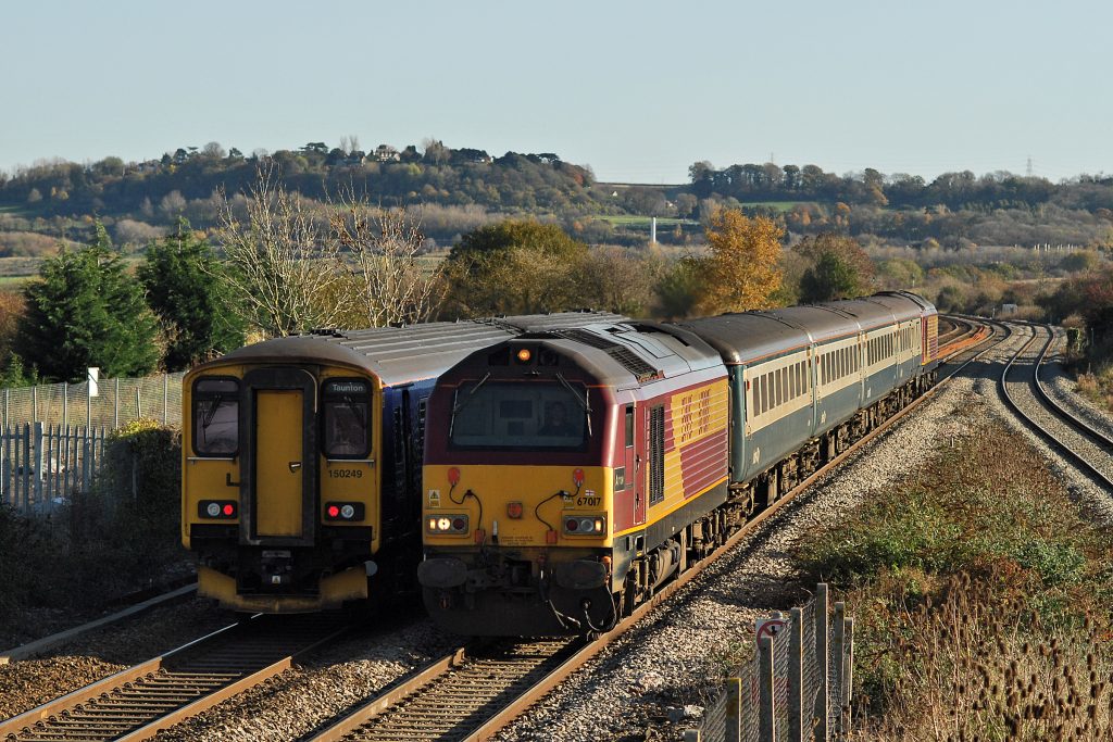 67017 & 67016 at rear are pictured passing Pilning with the 11.02 Taunton to Cardiff Central on 10th November 2010, as 150248 speeds through in the opposite direction with the 12.00 Cardiff to Taunton.  Locomotive haulage on these services finished after 12th November. The station footbridge from which the photograph was taken was partially dismantled during electrification of the route, and the down platform is no longer accessible.  Image credit: Stewart Jolly