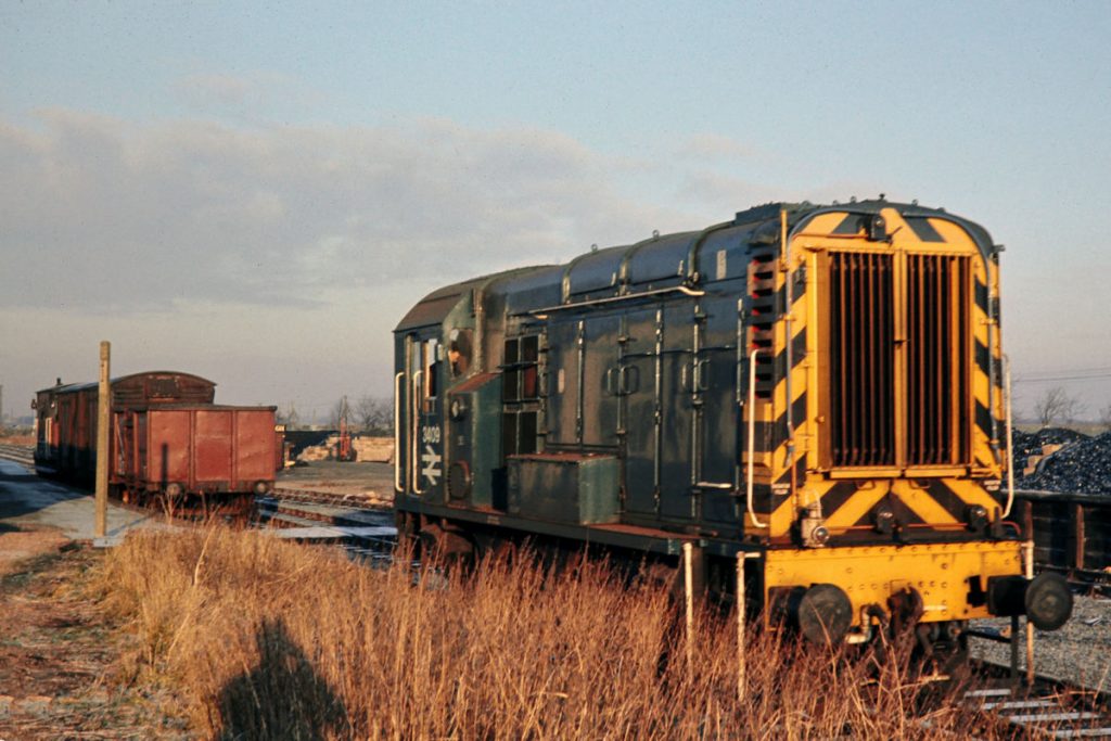 19701231-RAMSEY-NORTH-3409-08339-Running-Round-08 BR Class 08 No. 3409 has just arrived at Ramsey North on December 31st 1970 with 9B08, the 08.35 from Holme and has uncoupled and drawn forward, ready to run round its train. It will collect two empty 16-ton minerals from the adjacent sidings and then position the wagons it arrived with in the sidings, before heading back to Holme at 09.35. On arrival at Holme at 10.10 it will then head South at 10.50 to Connington as 9B25, arriving at 11.15 and finally head back to New England as 9B26, departing Connington at 12.35 and arriving in New England at 14.10. Photo: John Tolson, courtesy Trevor Davis.