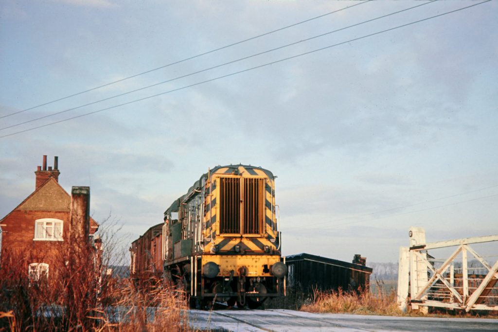 19701231-RAMSEY-St-MARYS-3409-08339-08 Long Drove crossing was the middle of the three gated crossing on the Ramsey North Branch and the flat, low-lying nature of the terrain can be seen in this view. With the crossing gates closed by the guard, Class 08 No. 3409 creeps over the B660 Holme to Ramsey St Mary's road with one mineral wagon and two vans that make up 9B08, the 08.35 from Holme to Ramsey North on December 31st 1970. Photo: John Tolson, courtesy Trevor Davis.