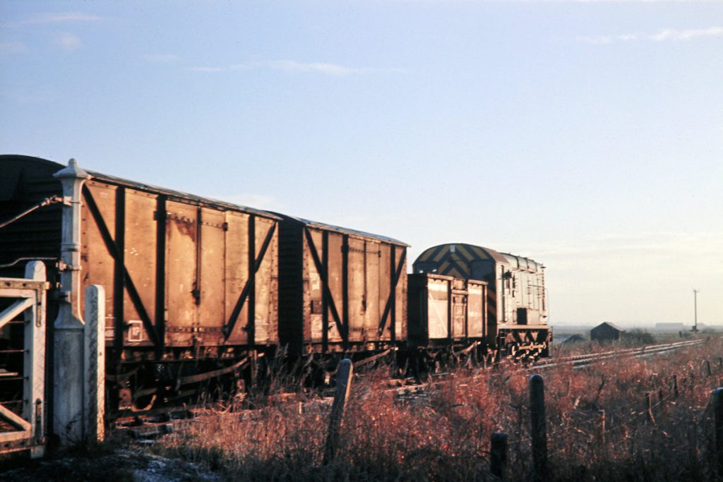 19701231-RAMSEY-St-MARYS-3409-08339-08 Drawing to a halt after crossing the road at Ramsey St Mary's on December 31st 1970, BR Class 08 No. 3409 (later 08 339) will pick up the guard and then head to Ramsey North on the final section of its run from Holme with 9B08. The ornate gate post on the left was a distinctive feature at St Mary's, being of cast iron rather than the usual heavy timber baulk. Photo: John Tolson, courtesy Trevor Davis.