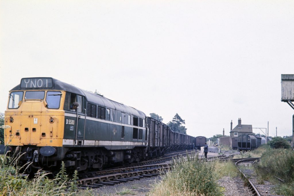 19700703-RYBURGH-D5580-06 Grain and agricultural traffic (e.g. seed potatoes and fertilisers) were important traffic on the railway through to the end of the 1970s. In this view of D5580 at Ryburgh on July 3rd 1970, grain wagons can be seen in the sidings to the right and the wagons at the station platform are about to be collected. Pick-up freight trains of this kind are simply no longer a part of the railway scene and everything is now transported by road, which is a great loss to the nation's overall effort to reduce carbon emissions. Photo: John Tolson, courtesy Trevor Davis.