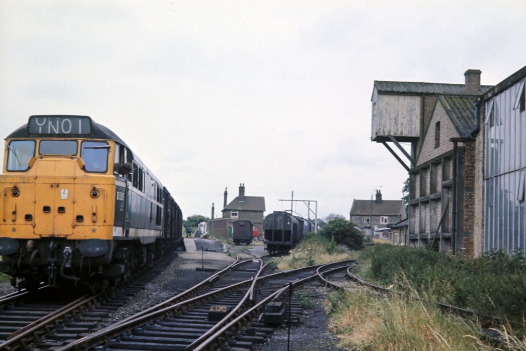 19700703-RYBURGH-D5580-06 The guard couples up wagons in the station platform to 8N01, the daily Wensum to Fakenham pick-up freight, on a dull Friday July 3rd 1970. Passenger services were withdrawn on 5th October 1964 when the Dereham-Wells service ceased and the line from Wells to Fakenham was closed. The line from Ryburgh to Fakenham was closed on 1st January 1980 and the final freight train to depart from Ryburgh was in August 1981. However, it is certainly not beyond the realms of possibility that the Mid-Norfolk Railway may once again bring trains into the town. Photo: John Tolson, courtesy Trevor Davis.