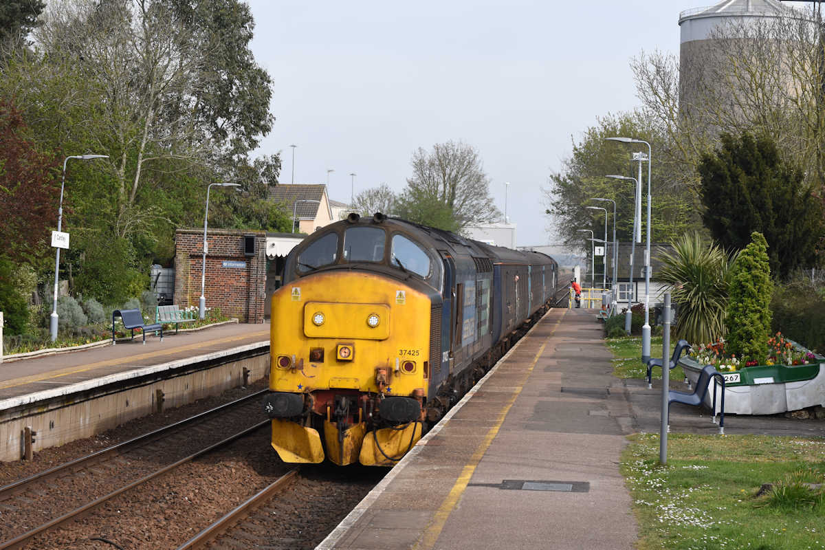 Cantley station is carefully tended by a group of local volunteers whose fine work can be seen. Drawing to a halt at the Up platform, with the Sugar Works to the right, 37425 and 37423 work the 15.48 Lowestoft to Norwich on  23rd April 2019. Photo: Alan Jones.