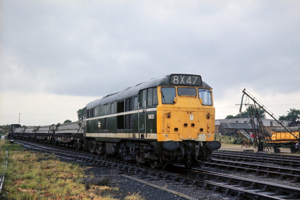 19700702-LENWADE-5621-16 Looking South at Lenwade towards Norwich City on July 2nd 1970, after a heavy downpour, Brush Type 2 No. 5621 prepares to return to Norwich with reinforced concrete beams on four bogie bolsters. The average speed of the journey was slow, with a 10 mph speed restriction round the Themelthorpe Curve and 25 mph all the way to Wroxham, meaning a journey from Norwich and back effectively took a whole shift. With a driver, second man, guard and signal man it is not difficult to see why the economics of this type of traffic were not strong. Photo: John Tolson, courtesy Trevor Davis.