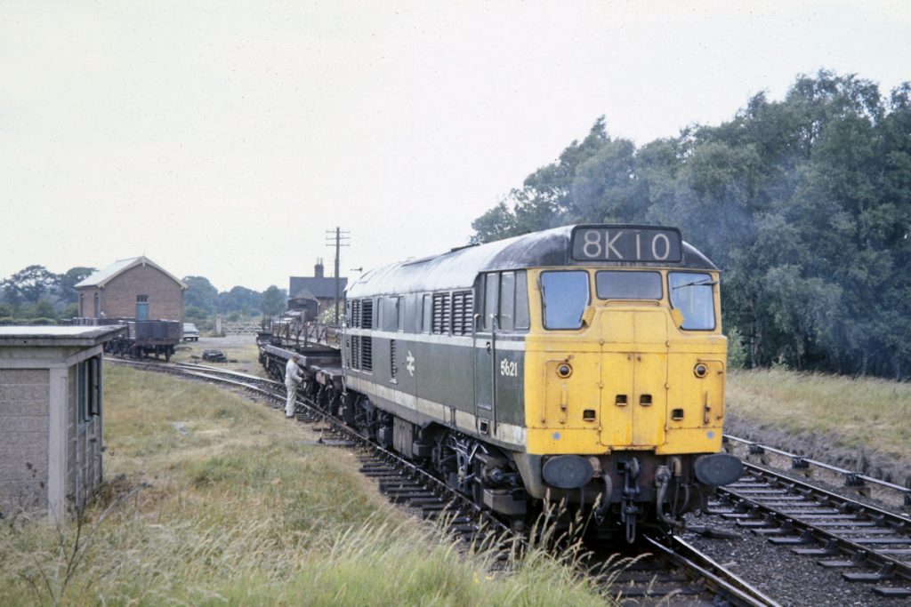19700702-LENWADE-5621-Draws-Steel-Griders-on-Bolsters-out-of-Sidings-JMT34942-John-Tolson-E4W The concrete works at Lenwade enjoyed a period of prosperity in the 1960s and early 70s due to the use of pre-cast steel and concrete building components, made at Lenwade, in the housing and North Sea Oil industries. Much of this traffic went by rail, but the line's fortunes declined when these components became too large to carry within the loading gauge and when it became apparent that they had poor performance in fire conditions. The last train ran in 1981, but the line from Aylsham to Wroxham is now part of the Bure Valley Railway. On July 2nd, 1970 Brush Type 2 No. 5621 draws steel girders out of the small yard and into the concrete works. Photo: John Tolson, courtesy Trevor Davis.