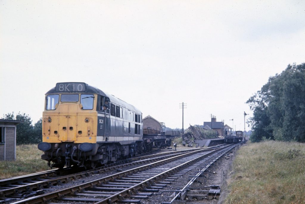 19700702-LENWADE-5621-Draws-Steel-Griders-on-Bolsters-out-of-Sidings-JMT34943-John-Tolson-E2W A delightful scene captured by John Tolson at Lenwade station on July 2nd 1970, in which Brush Type 2 No. 5621 draws a short rake of bolsters out of the station yard to run down to the Concrete Works behind the photographer. Although the passenger service was withdrawn in February 1959, Norwich City station continued as the city's main coal depot until replaced by one at Victoria in 1969, at which time the line was cut back to Drayton to serve the gravel depot and then subsequently cut back to Lenwade. Photo: John Tolson, courtesy Trevor Davis.