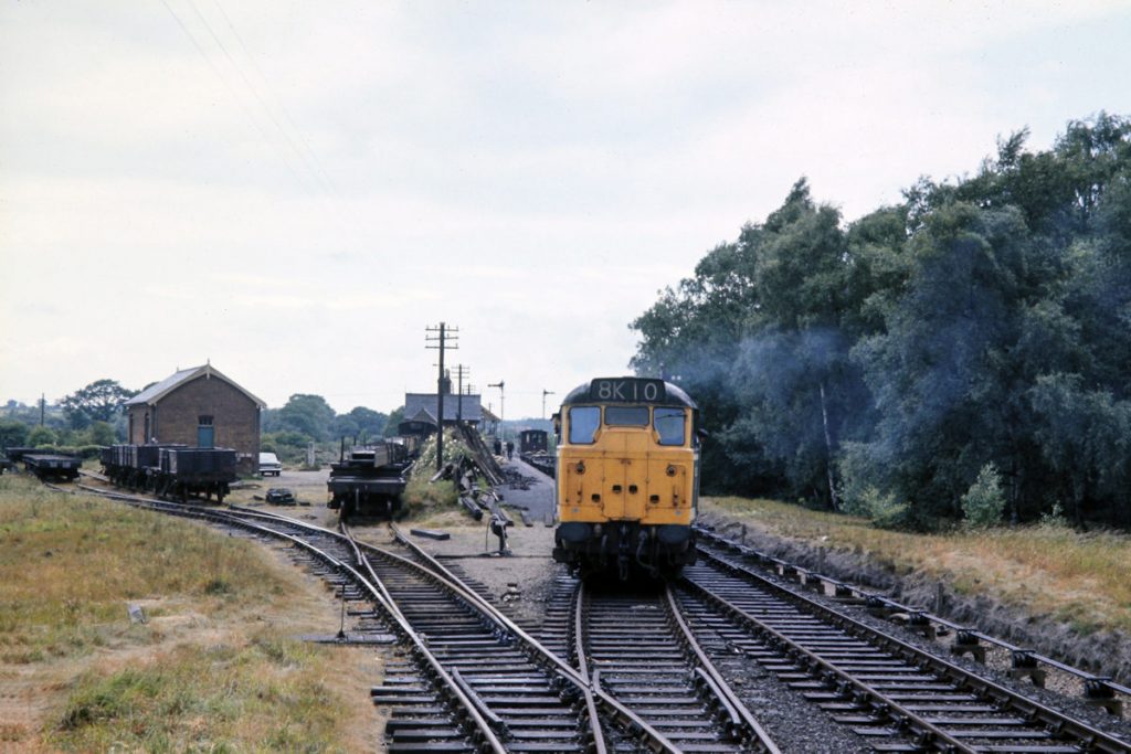 19700702-LENWADE-5621-Runs-Light-Engine-into-Sidings-after-arrival-from-Norwich-JMT34941-John-Tolson-E4W On Thursday 2nd July, 1970 Brush Type 2 No. 5621 has just arrived at Lenwade and dropped off its empty bolsters, having travelled from Norwich via the Themelthorpe Curve. This connection was opened in September 1960 to join the former Wroxhan-County School line to the M&GN line South from Melton Constable. Between closure of the M&GN system in February 1959 and opening of the Themelthorpe curve, trains for the Norwich City line had to travel up to Cromer then round through Sheringham and Melton Constable - a route so circuitous they were known to railwaymen by the term 'Round the World'! Note the unusual catch points in the pointwork at the exit from the sidings. Photo: John Tolson, courtesy Trevor Davis.