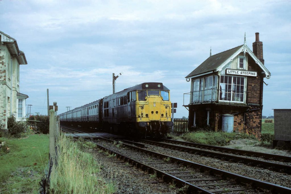 19850831-LITTLE-STEEPING-31247-13-30-SO-Skegness-Leeds-Alan-Jones-EW The friendly owners of the former station house at Little Steeping offered an opportunity to photograph 31247 from their garden as it worked the 13.30 (SO) Skegness to Leeds on August 31st 1985. Little Steeping Box became increasingly unsafe as it slid further into the marsh, but all the while it retained its delightful Great Northern characteristics, including original nameboards, bargeboards and both finials. Curiously, although the lean of the Box is pronounced, the chimney seems to be leaning in the opposite direction! Photo: Alan Jones.