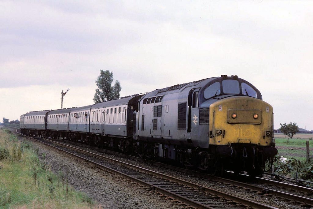 19870926-SIBSEY-37239-Grantham-Skegness-Additional-Alan-Jones-EW One of the early 1980s cost cutting measures implemented by BR on the Skegness line was the singling of the line between Hubbert's Bridge and Sibsey, with a passing loop at Boston station. The Down line was retained East of Boston, but in order to make use of the existing trailing crossover at Sibsey to regain the double track the Down line had to be slewed, resulting in an awkward 20 mph speed restriction. Clanking over the pointwork, a very grubby 37239 works a Grantham to Skegness additional on September 26th 1987. Photo: Alan Jones.