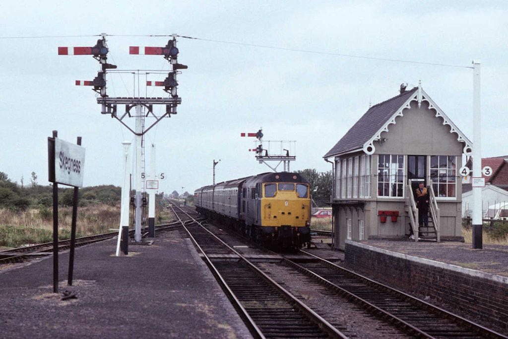 19890909-SKEGNESS-31422-07-30-SO-ex-Birmingham-New-Street-2-Alan-Jones-EW For most of the year Skegness station saw little activity, with little more than a two-car DMU every two hours. But on Summer Saturdays it came to life with locomotive-hauled trains, propelling movements, running round and the rusty outer platform lines brought into use. The shunter has just finished a cup of tea in the signal box and heads down to the platform to meet 31422 arriving with the 07.30 (SO) from Birmingham New Street on September 9th 1989. He will join the train at the rear and wave it out of the station into the carriage sidings, then run the locomotive round and wave the train back in as it reverses into the station. Photo: Alan Jones.