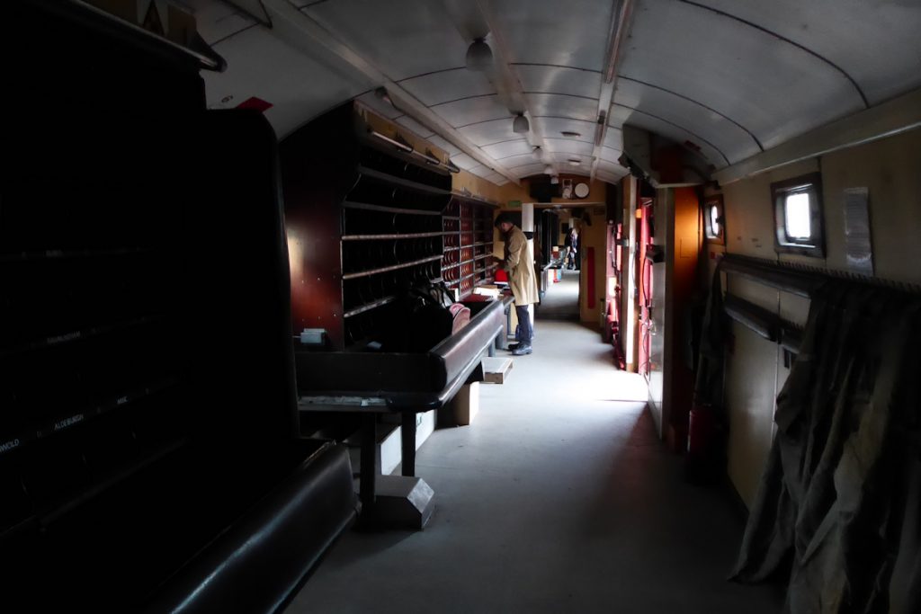 20240928-WANSFORD-80337-Brian-White-and-80327-George-James-19 An atmospheric view down the two TPO carriages 80337 and 80327 on the Nene Valley Railway's 'Night Mail' special on Saturday 28th September 2024. The light on the side of the newspaper (or packet) frame in position 7 is coming from the window of the door in position 6, and the folding table that drops down in front of this door can be seen stowed upright at the end of the Newspaper rack in position 5. Photo: Alan Jones.