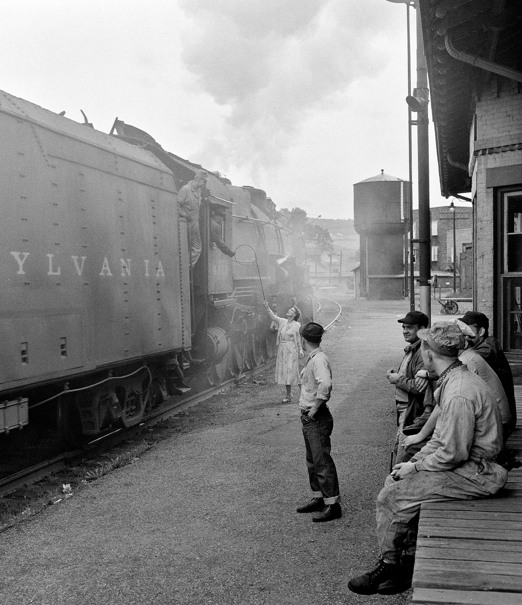 In this image by Jim Shaughnessy, Louise Overhiser, the block operator at the Pennsylvania’s Watkins Glen, New York, station hands orders up to the engineer of a big 386,000-pound I-1 class 2–10–0 #4512, as it thunders past her with a string of empty coal hopper cars. Nearby, the crew of an Elmira Branch local freight wait for the passing train to clear so they can come out of a side track and continue north. Image shot by Shaughnessy, circa 1955.